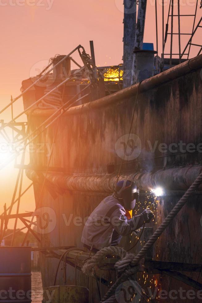 vue latérale du soudeur en vêtements de protection sur la plate-forme soude la paroi métallique de l'ancien bateau de pêche dans la zone du chantier naval au coucher du soleil dans un cadre vertical photo