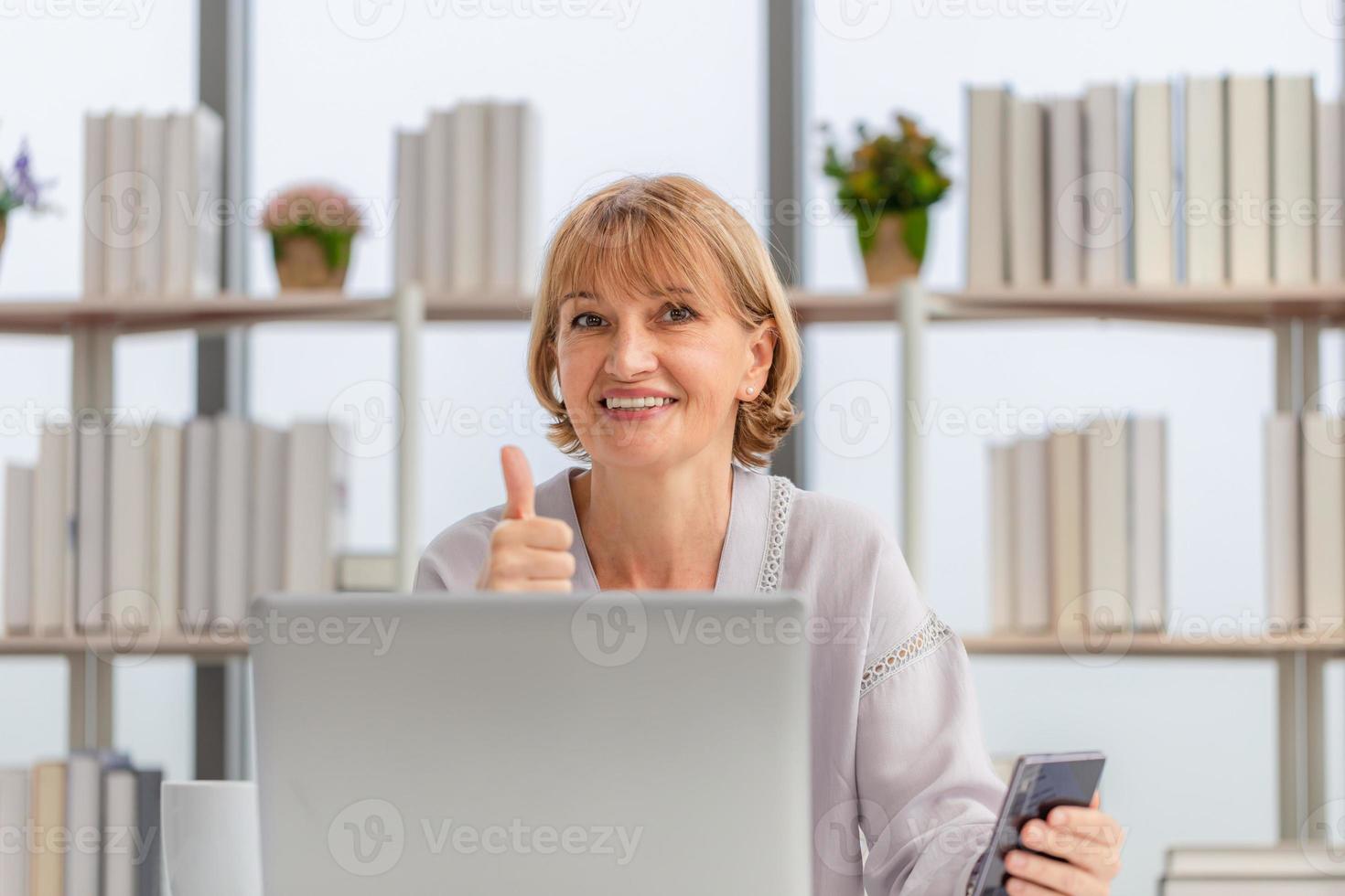 portrait d'une femme utilisant un ordinateur portable et un smartphone à la maison et montrant les pouces vers le haut, une femme mûre heureuse dans le salon avec un ordinateur portable parlant un appel vidéo sur un gadget informatique moderne photo