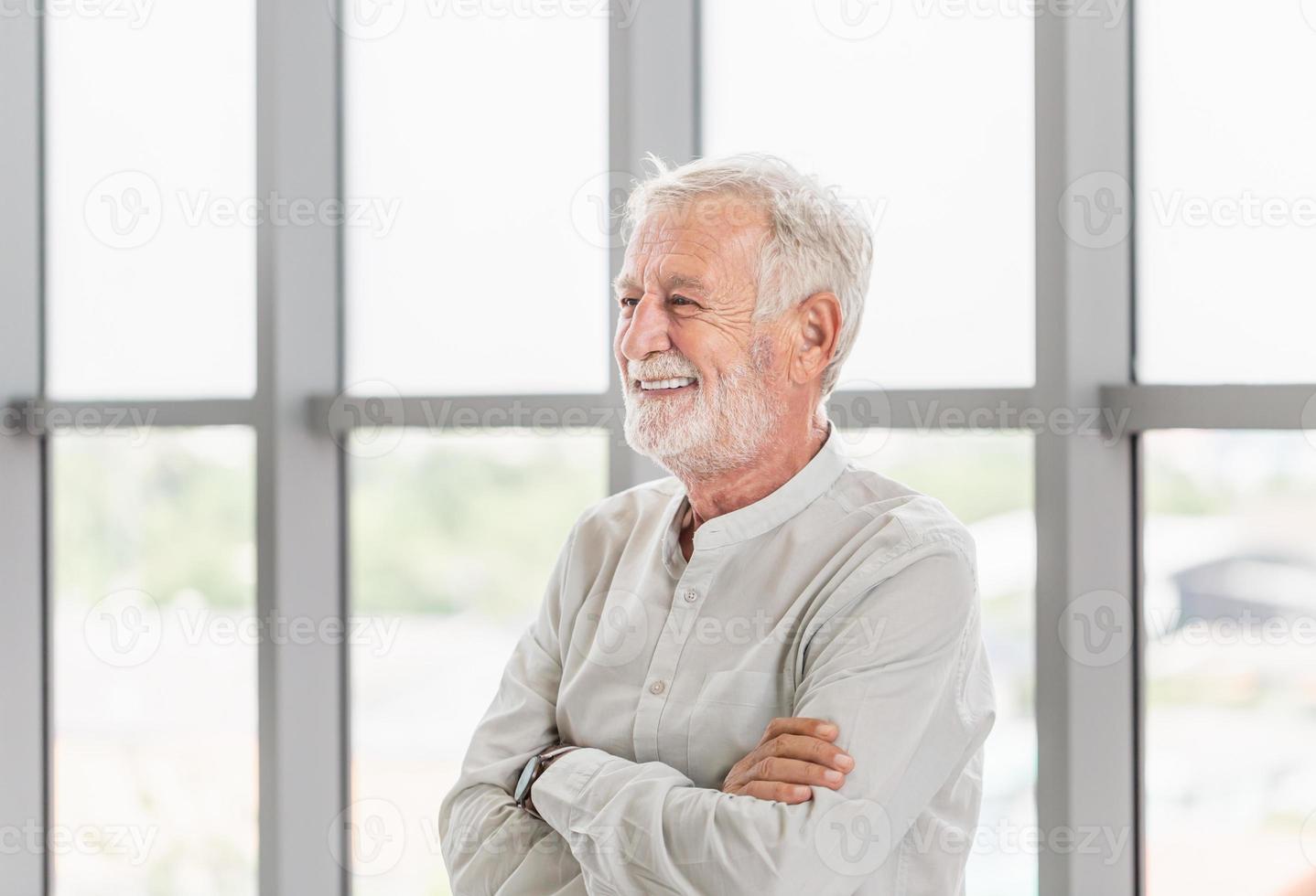Beau vieil homme de race blanche senior souriant avec les bras croisés, heureux vieil homme debout près de la fenêtre photo