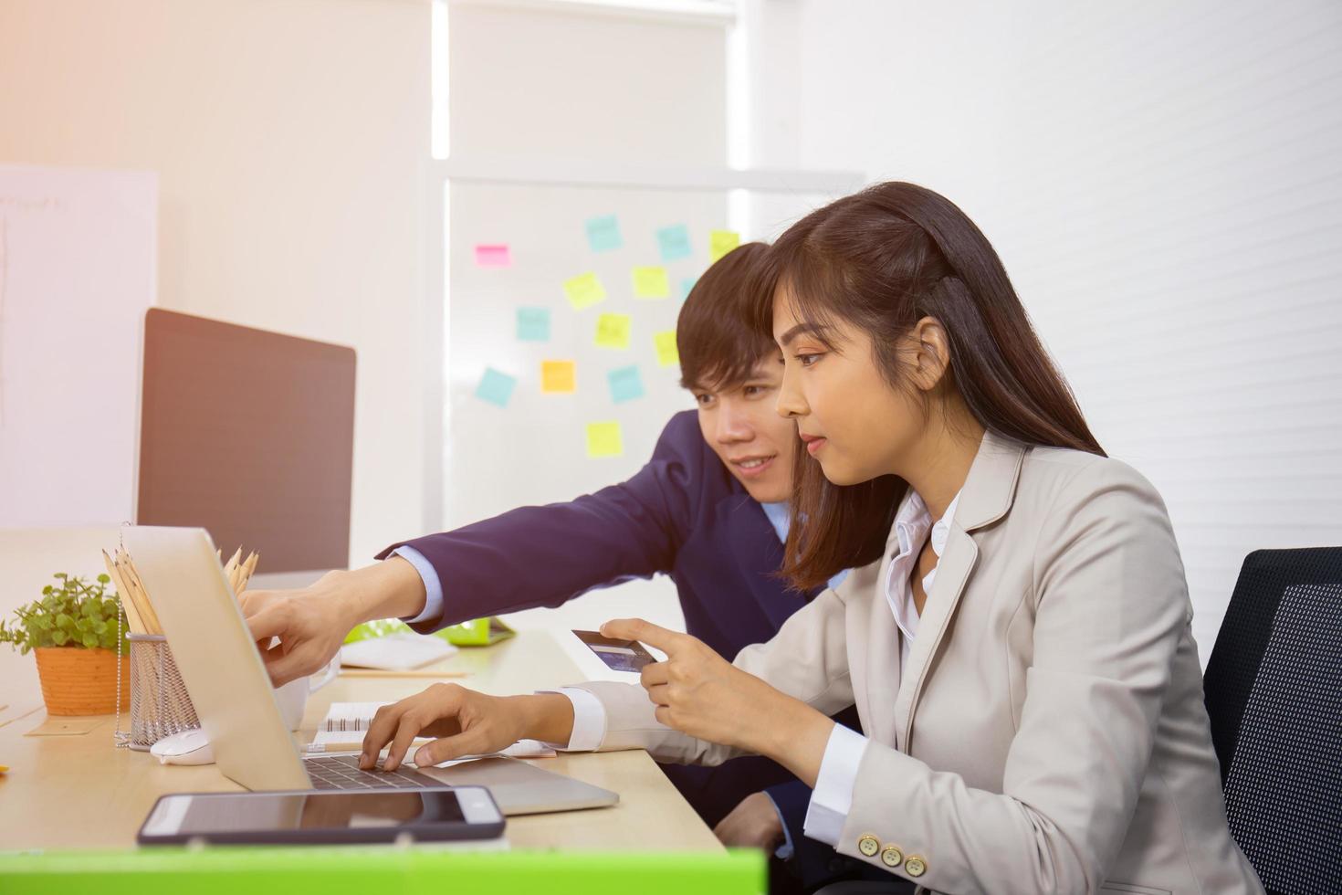 une jeune femme d'affaires asiatique professionnelle qui utilise un ordinateur portable et une carte de crédit pour payer en ligne tout en étant assise à son bureau, avec un ami masculin à côté de lui. photo