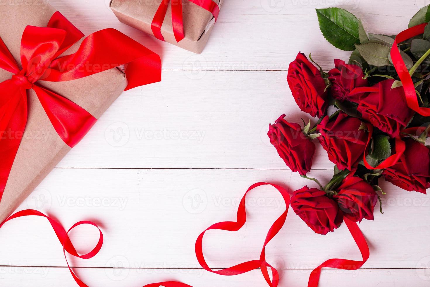 ruban rouge en forme de coeur, bouquet de roses et coffrets cadeaux enveloppés dans du papier kraft sur une table en bois blanche. photo