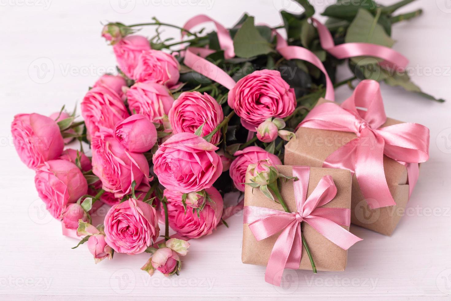 des roses rose pastel fraîches et des coffrets cadeaux enveloppés dans du papier kraft avec des rubans sur une table en bois blanche. photo