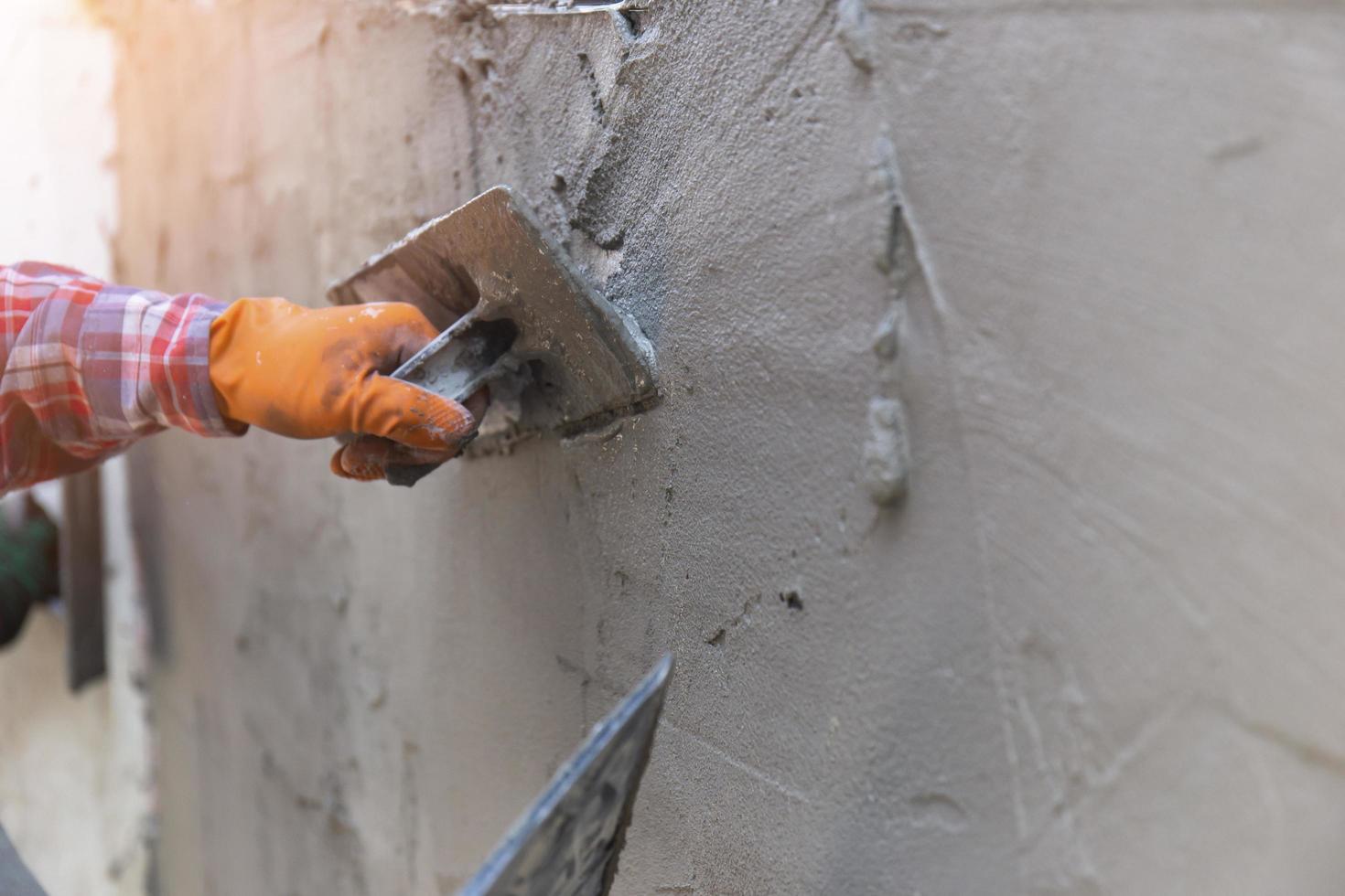 les mains d'un ouvrier qui plâtre de près portent des gants en caoutchouc orange pour empêcher le ciment de se mordre les mains, construisant les murs de la maison et ayant de belles lumières orange. photo