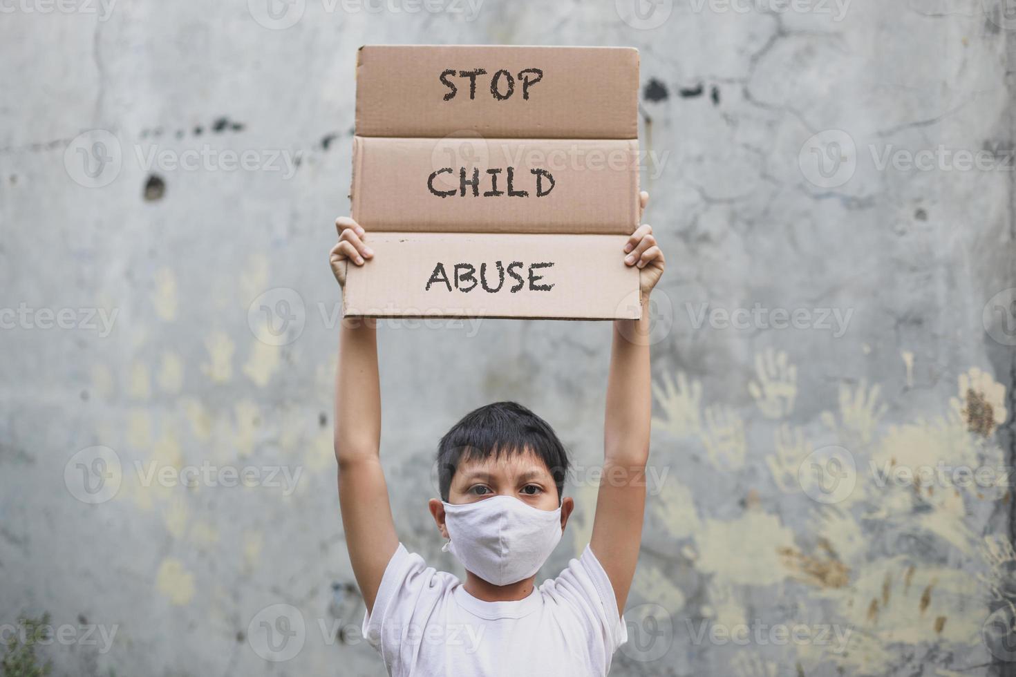 garçon asiatique en masque médical levant le tableau des lettres dit stop à la campagne contre la maltraitance des enfants avec empreinte de la main dans le fond du mur de ciment photo