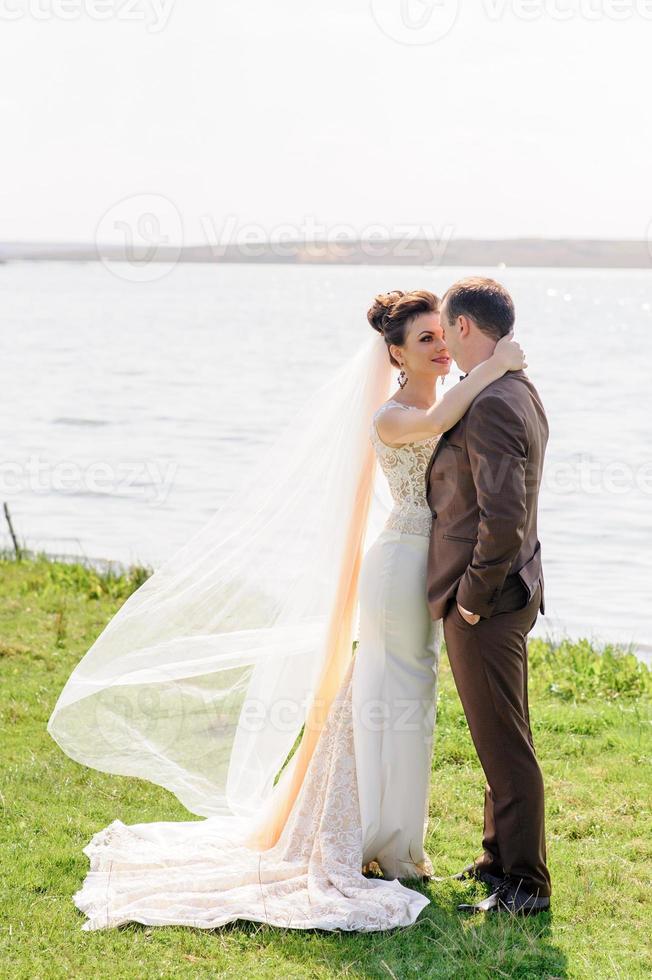 la mariée et le marié s'embrassent près de l'étang dans la nature. le vent soulève un léger voile. photo