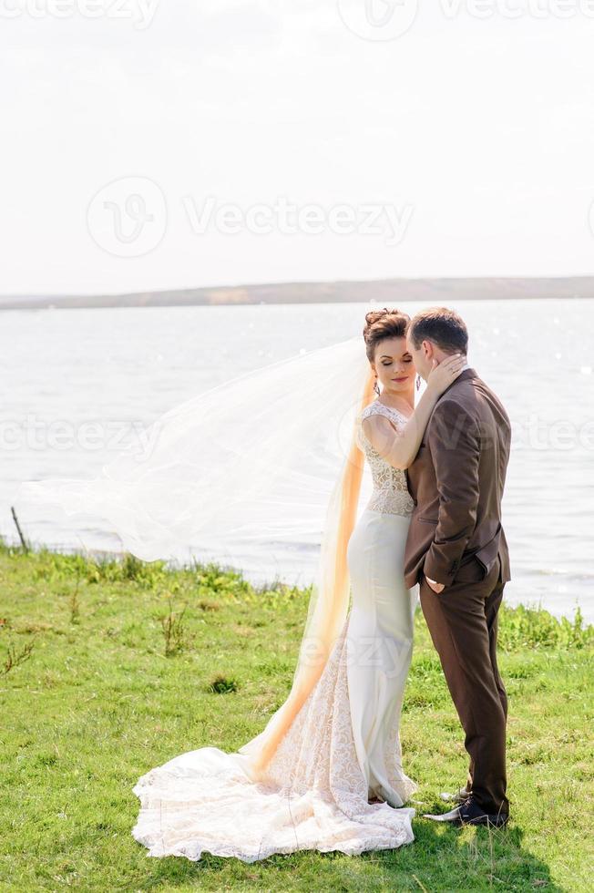 la mariée et le marié s'embrassent près de l'étang dans la nature. le vent soulève un léger voile. photo