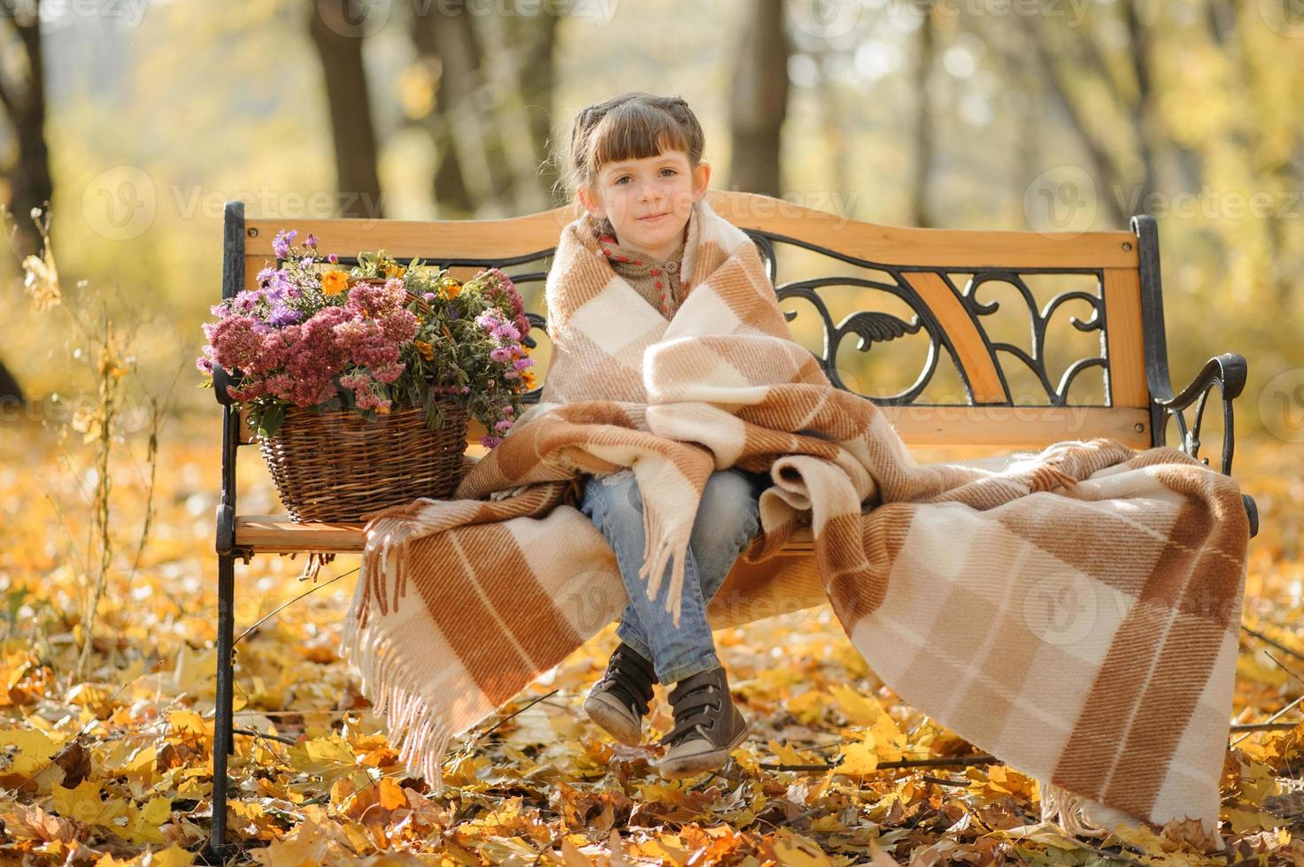petite fille mignonne est assise sur un banc dans le parc d'automne. la jeune fille s'abrita pour se réchauffer. temps de l'automne. photo