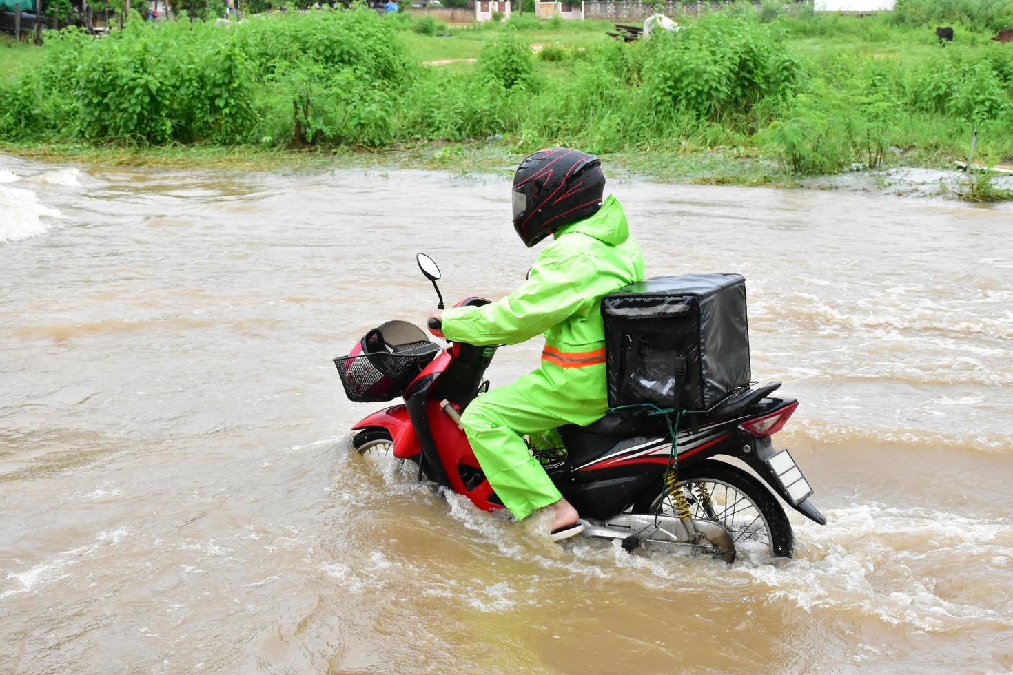 livraison homme asiatique portant un uniforme vert, chevauchant et livrant de la nourriture au client avec une boîte de nourriture derrière dans la rue inondée, livraison express de nourriture et concept d'achat en ligne dans la situation d'inondation. photo