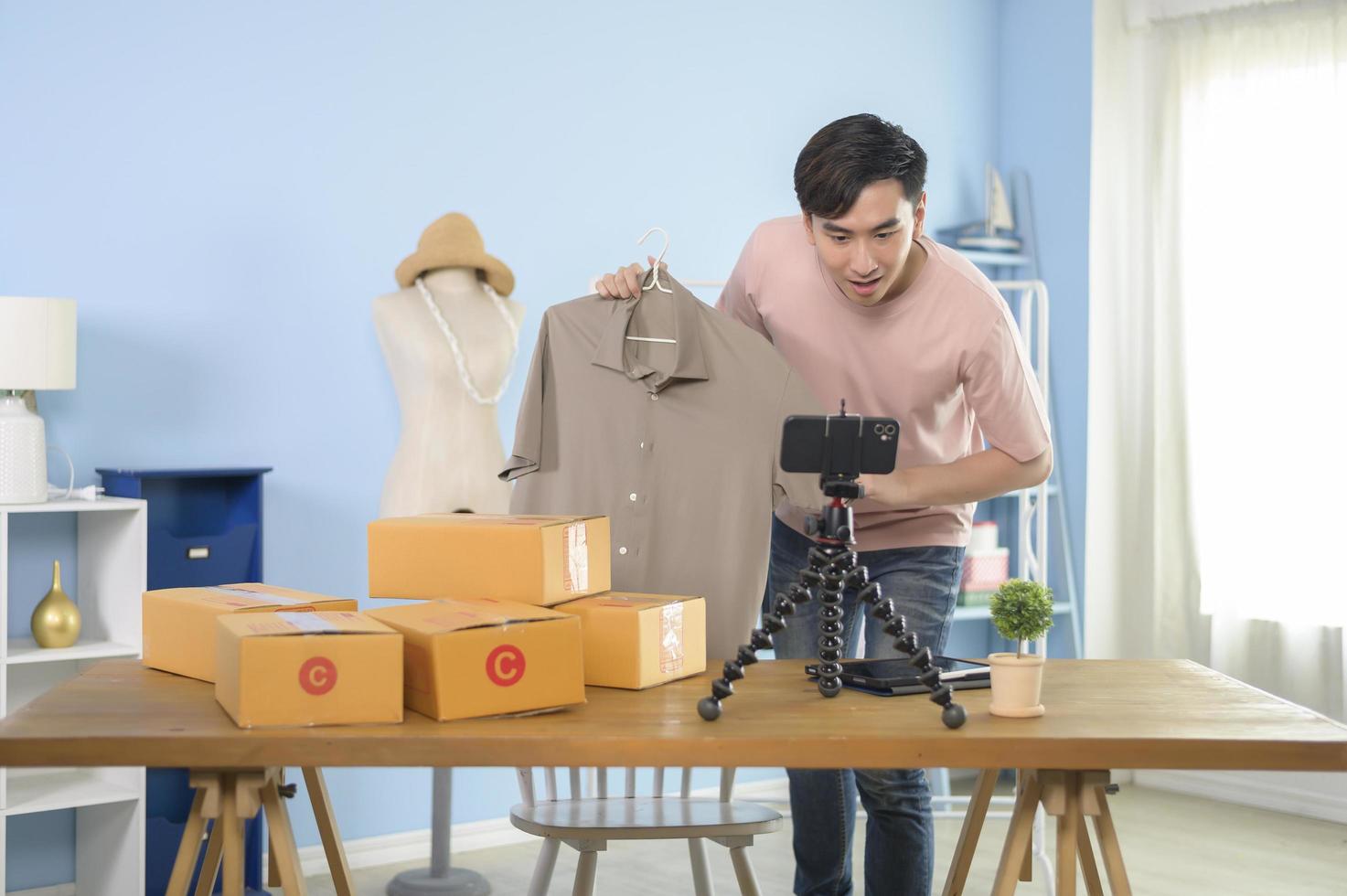 un homme asiatique montre des vêtements devant la diffusion en direct d'un smartphone dans sa boutique. concept d'entreprise en ligne de technologie. photo