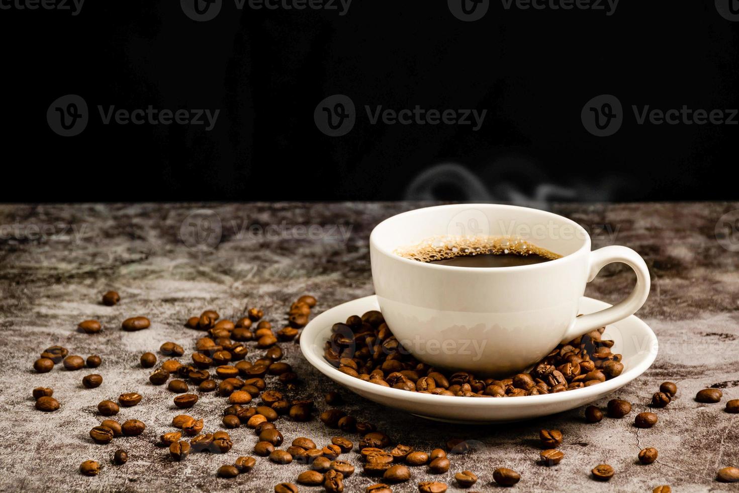 un gros plan de café chaud dans une tasse blanche est placé sur une table de sol en ciment, beaucoup de grains de café torréfiés sont dans une soucoupe de tasse à café, et autour, de la fumée et de l'arôme s'échappent de la tasse. fond noir et flou photo