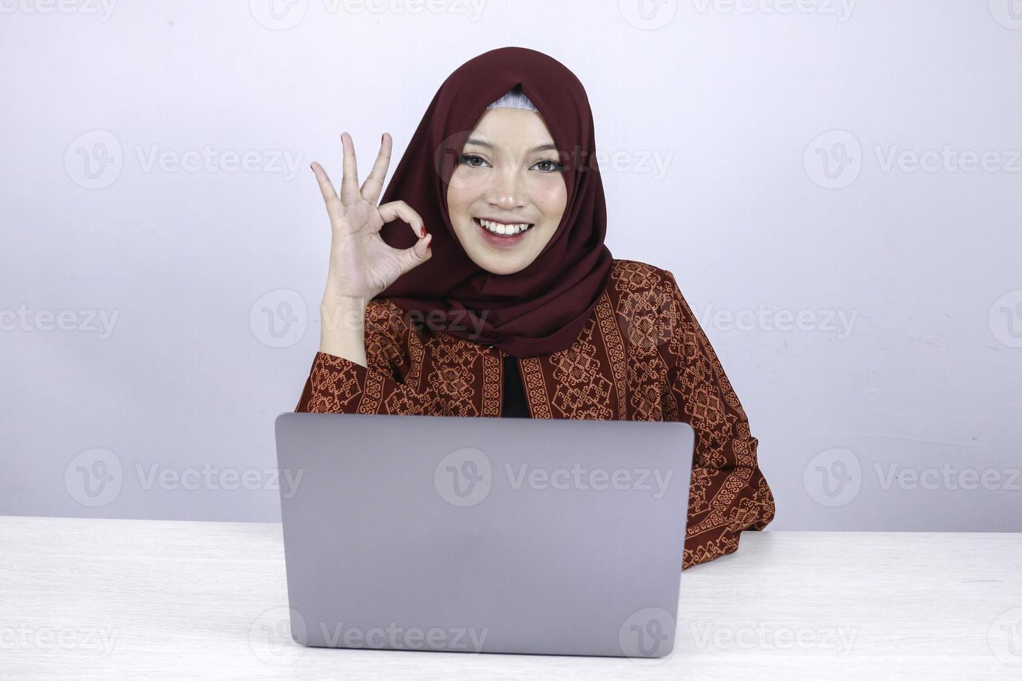 la jeune femme islamique asiatique est souriante et heureuse lorsqu'elle travaille sur un ordinateur portable avec le signe de la main ok. photo
