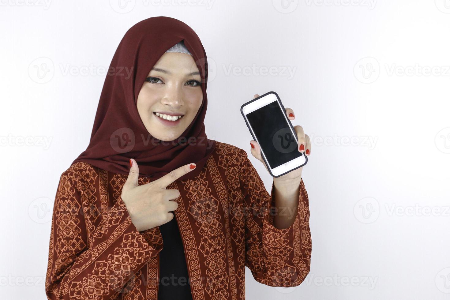 jeune femme islamique asiatique sourit en pointant sur un smartphone debout sur fond blanc. photo