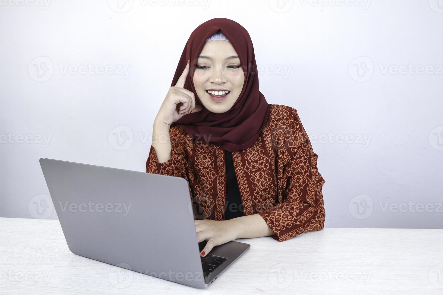 jeune femme islamique asiatique sourit avec un geste de pensée regardant sur l'espace vide à l'avant de l'ordinateur portable photo