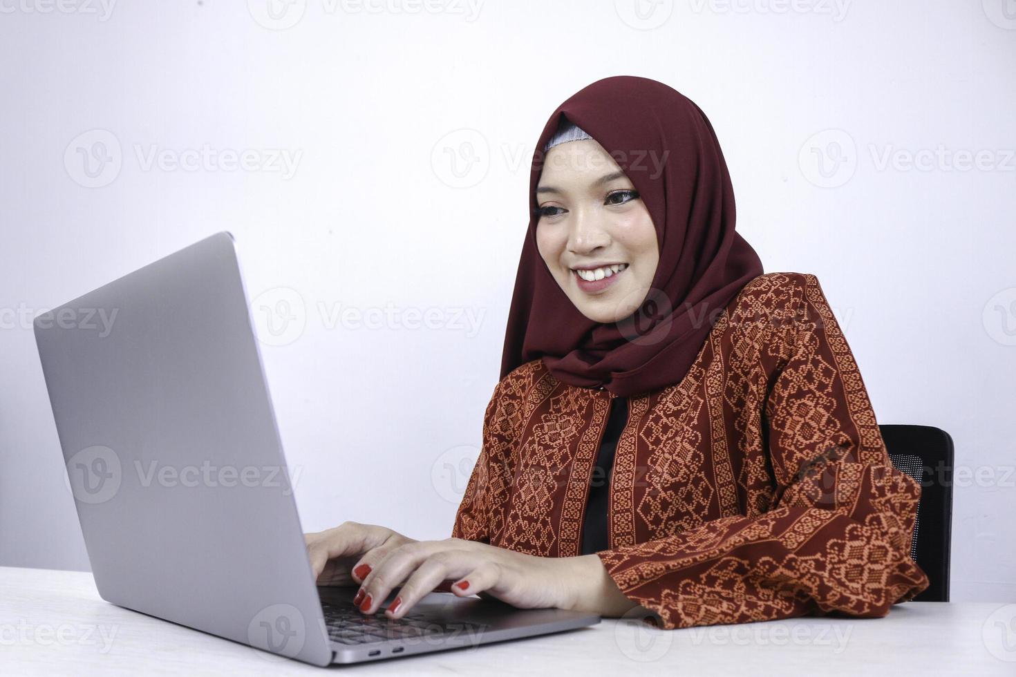 jeune femme islamique asiatique est assise et sourit en travaillant sur un ordinateur portable sur fond blanc. photo