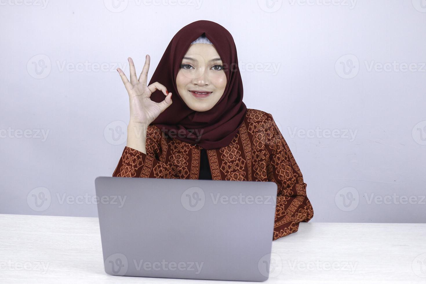 la jeune femme islamique asiatique est souriante et heureuse lorsqu'elle travaille sur un ordinateur portable avec le signe de la main ok. photo