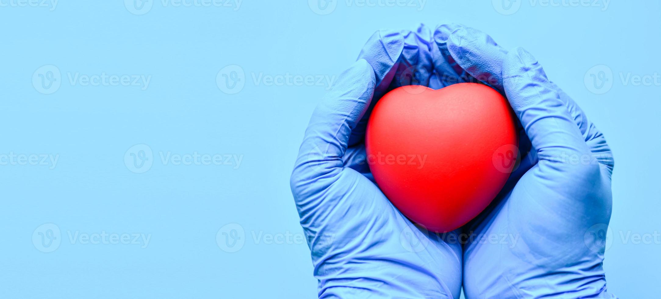 les deux mains portaient des gants et tenaient un cœur rouge. concept world heart foundation organise la journée mondiale du coeur, copiez l'espace à gauche pour la conception ou le texte, gros plan, arrière-plan bleu flou photo