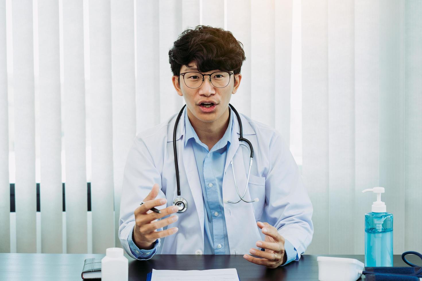 un médecin asiatique portant des lunettes et des uniformes blancs avec un stéthoscope fournit des conseils en ligne aux patients pendant l'épidémie de virus en gardant une distance sociale. photo
