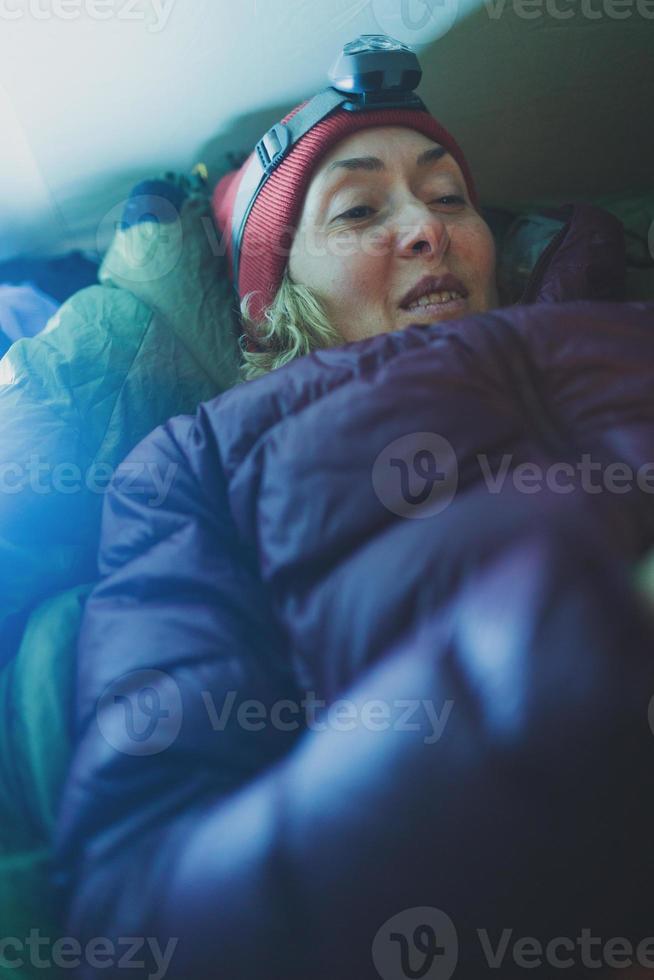 une femme avec une lampe frontale se trouve dans un sac de couchage dans une tente et tient un smartphone photo