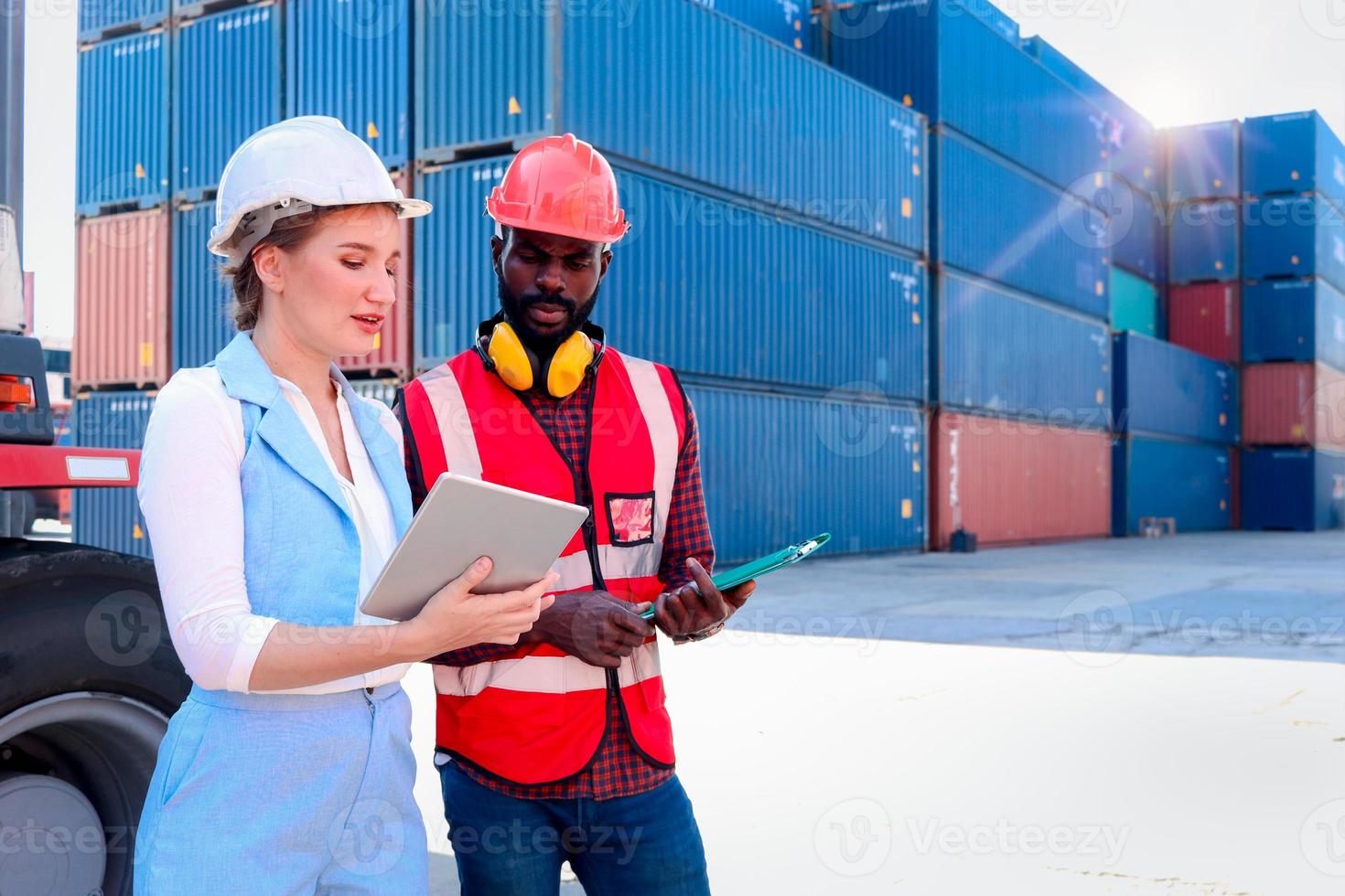 deux travailleurs portant un gilet de sécurité et un casque discutant dans un chantier de conteneurs de fret logistique. ingénieur afro-américain parlant avec une belle jeune femme patron aux cheveux blonds sur le lieu de travail. photo