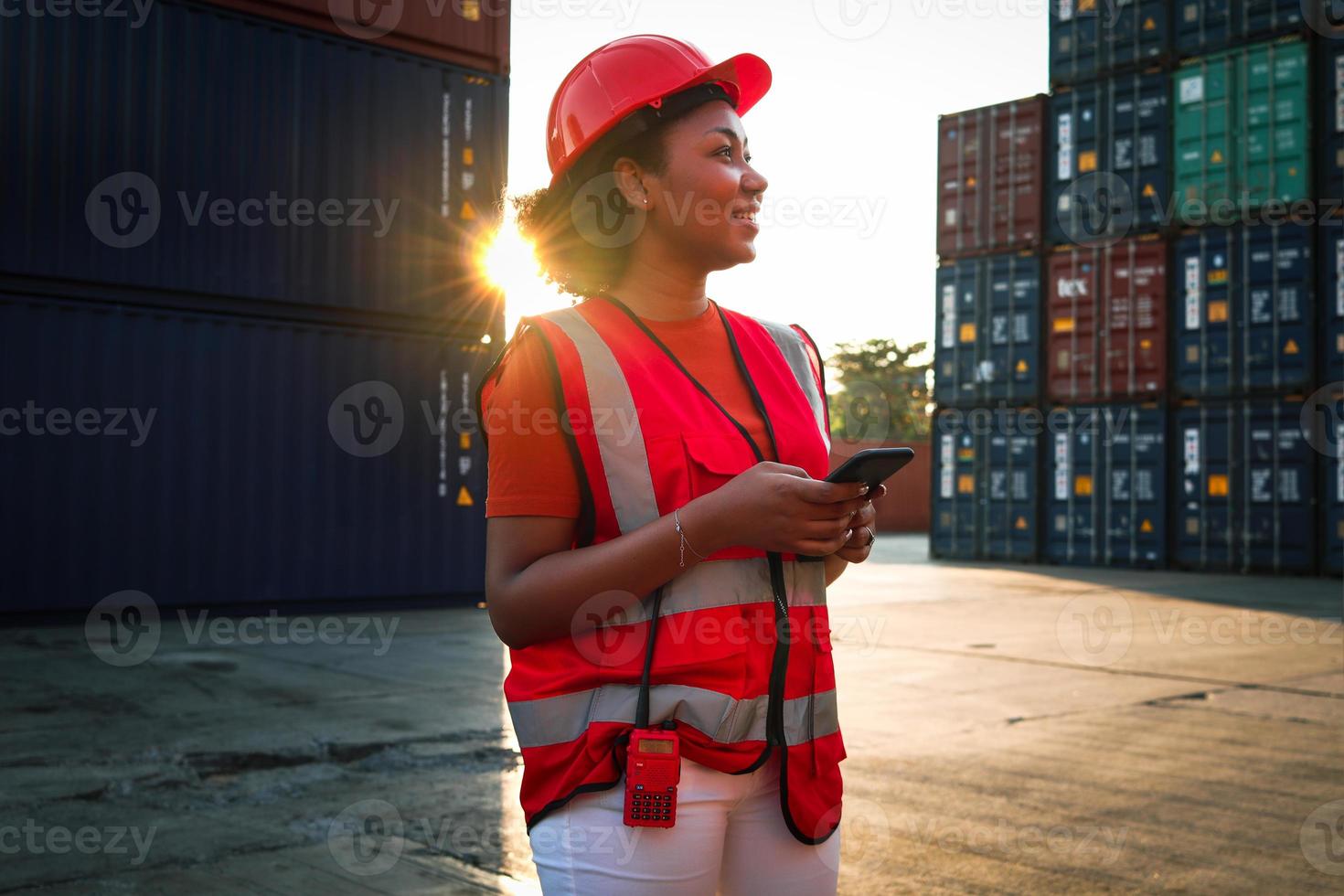 femme ingénieure afro-américaine aux cheveux bouclés portant un gilet de sécurité et un casque, tenant un téléphone portable au crépuscule au coucher du soleil dans la cour de conteneurs de fret d'expédition logistique. photo