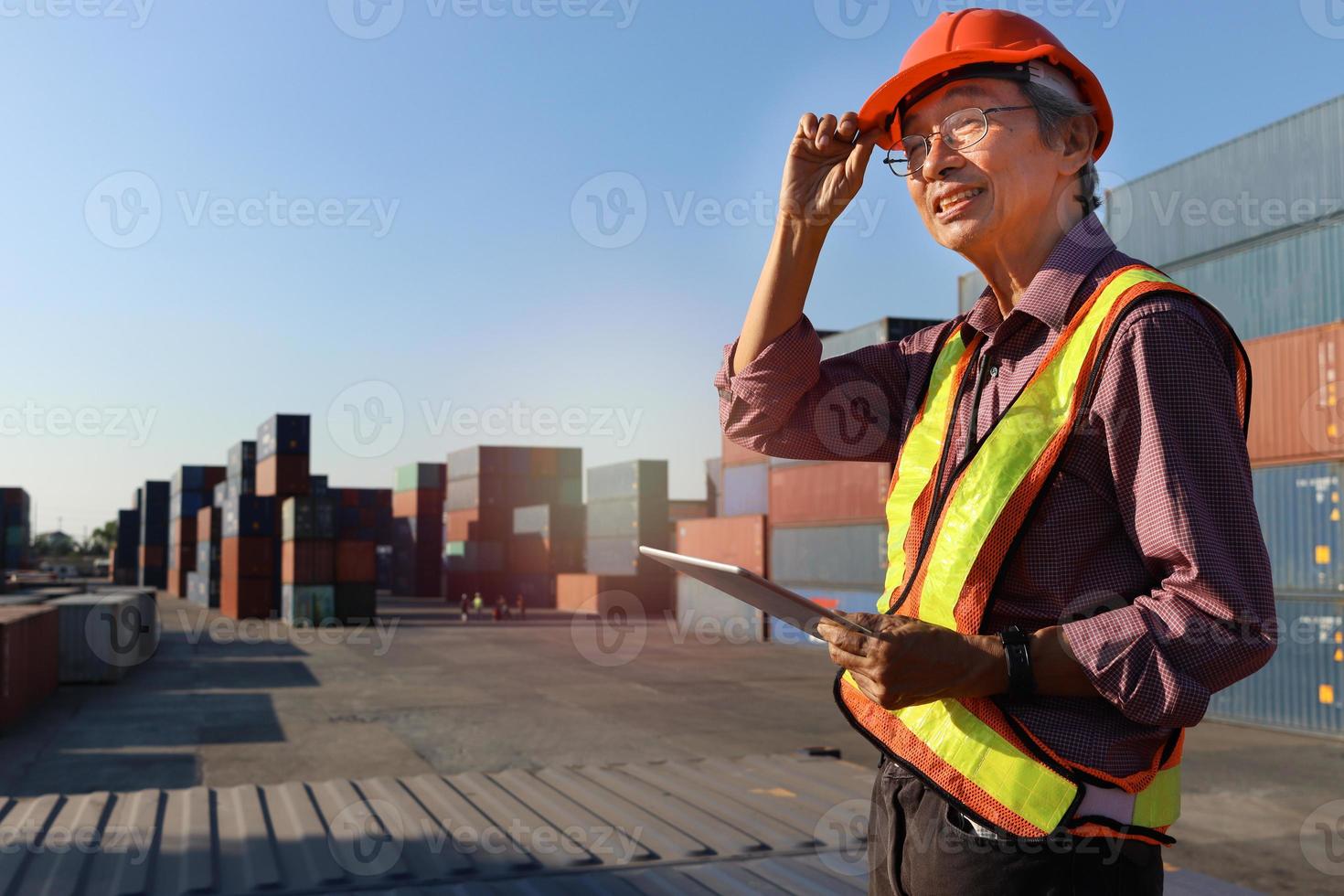 un ingénieur asiatique senior âgé portant un gilet de sécurité et un casque debout et tenant une tablette numérique dans la cour des conteneurs de fret d'expédition. personnes âgées au concept de lieu de travail photo