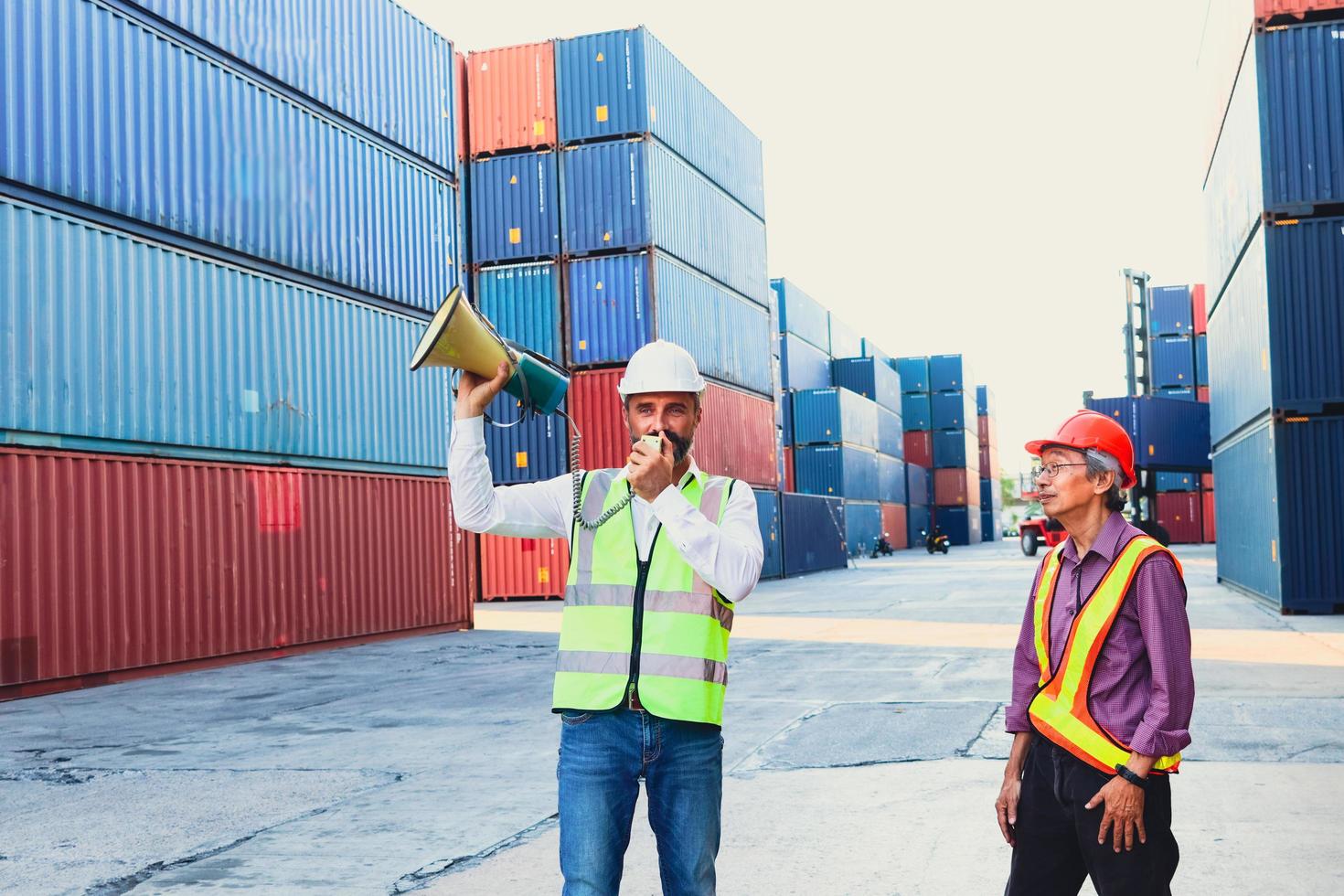 deux travailleurs avec gilet de sécurité et casque travaillant dans la cour de conteneurs de fret d'expédition logistique, le patron de l'ingénieur tenant un haut-parleur mégaphone pour communiquer avec le personnel asiatique âgé sérieux. photo