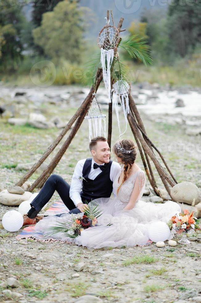 couple attrayant jeunes mariés, moment heureux et joyeux. homme et femme en vêtements de fête s'assoient sur les pierres près de la décoration de mariage dans un style bohème. cérémonie en plein air. photo