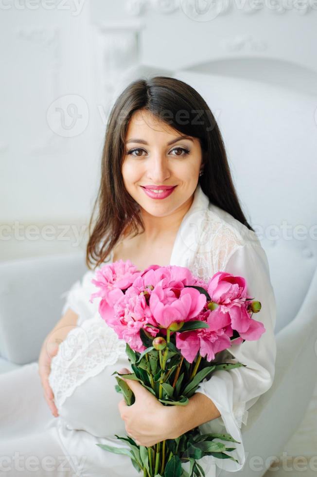 photo en gros plan d'une femme enceinte avec un ventre nu tenant un bébé chaussons blancs et fleurs roses sur un lit blanc