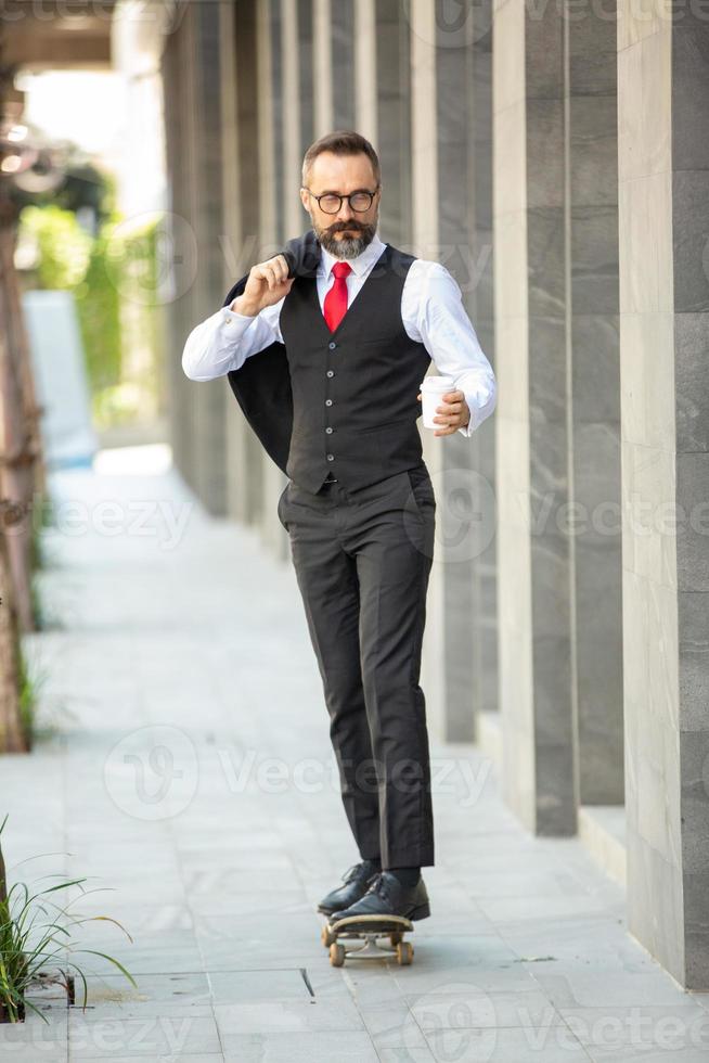 homme d'affaires hipster en costume debout sur une planche à roulettes avec une tasse de café près du bureau en plein air photo