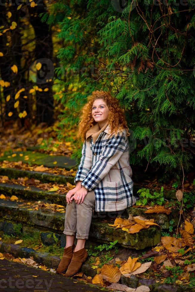 belle fille rousse aux cheveux bouclés et aux yeux bleus. la fille porte une veste à carreaux. la fille est assise sur un banc. photo