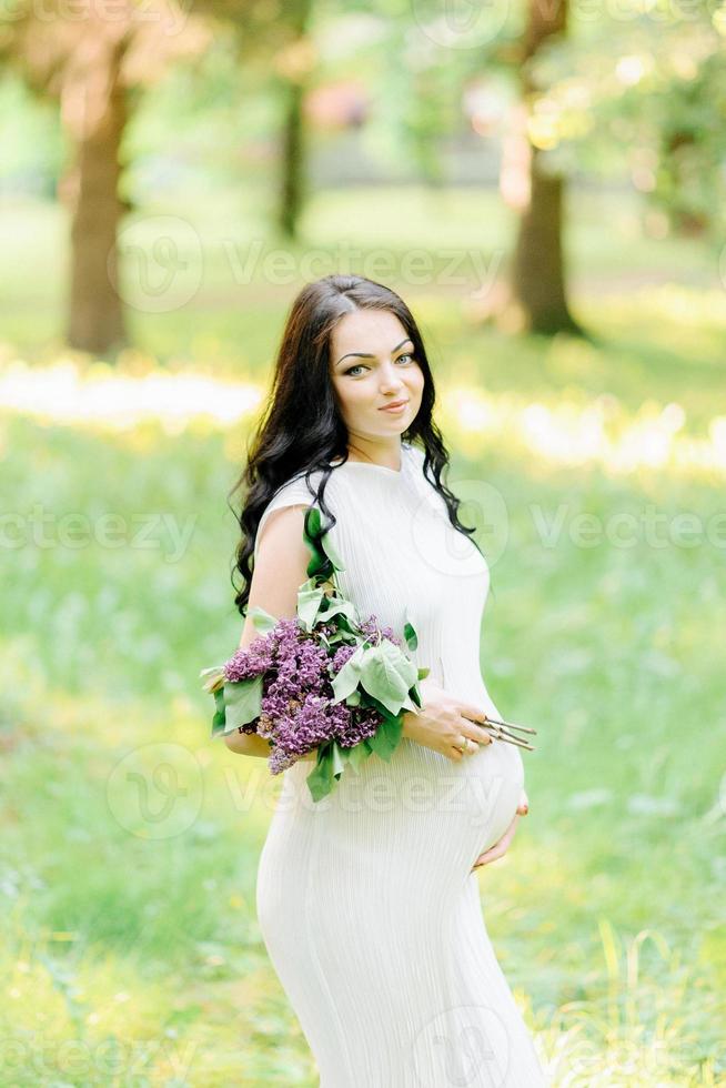 jeune femme enceinte au printemps jardin fleuri lilas. look romantique avec chapeau de paille. femme caucasienne aux longs cheveux bruns. concept de nouvelle vie de la nature et de l'homme. attente de bébé photo