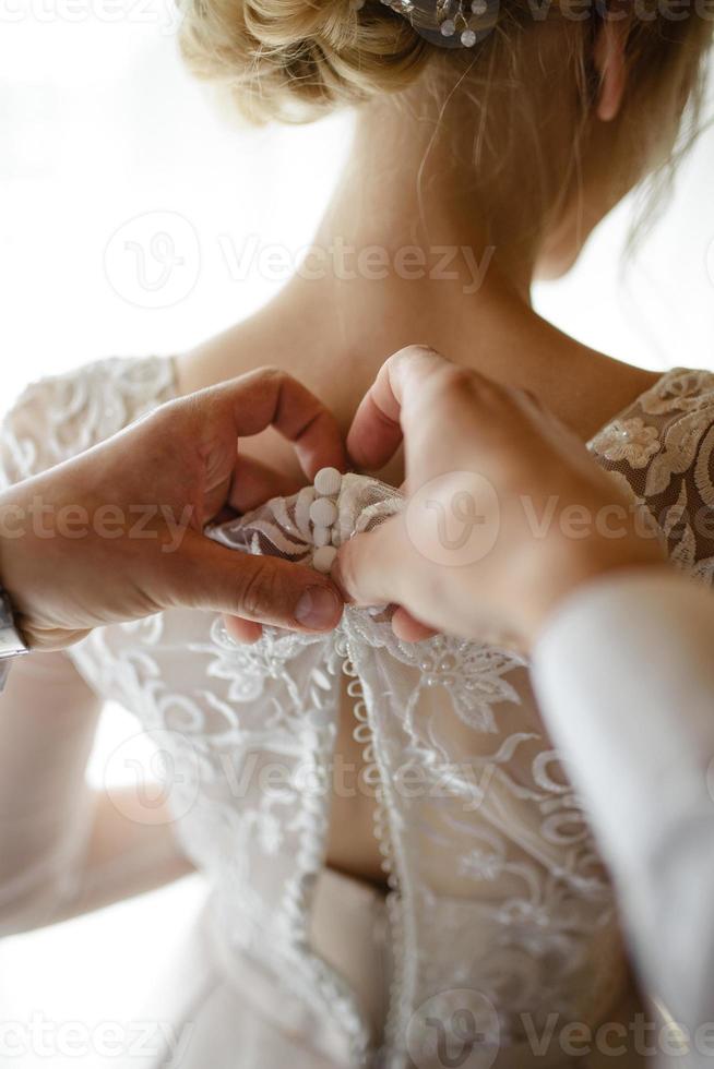 magnifique mariée blonde vêtue d'une luxueuse robe blanche se prépare pour le mariage. préparatifs du matin. une femme met une robe. le marié aide la mariée. photo