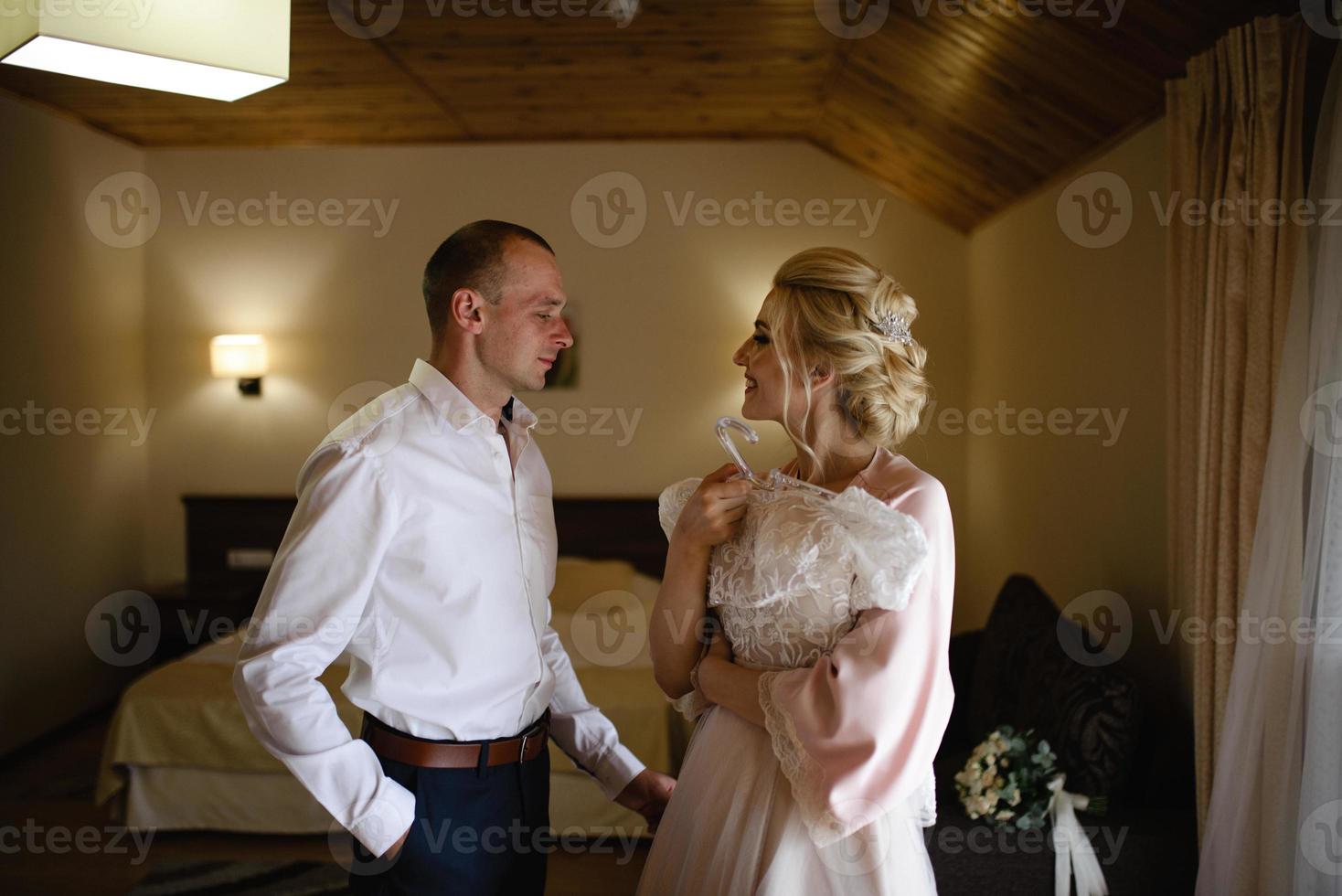 magnifique mariée blonde vêtue d'une luxueuse robe blanche se prépare pour le mariage. préparatifs du matin. une femme met une robe. le marié aide la mariée. photo