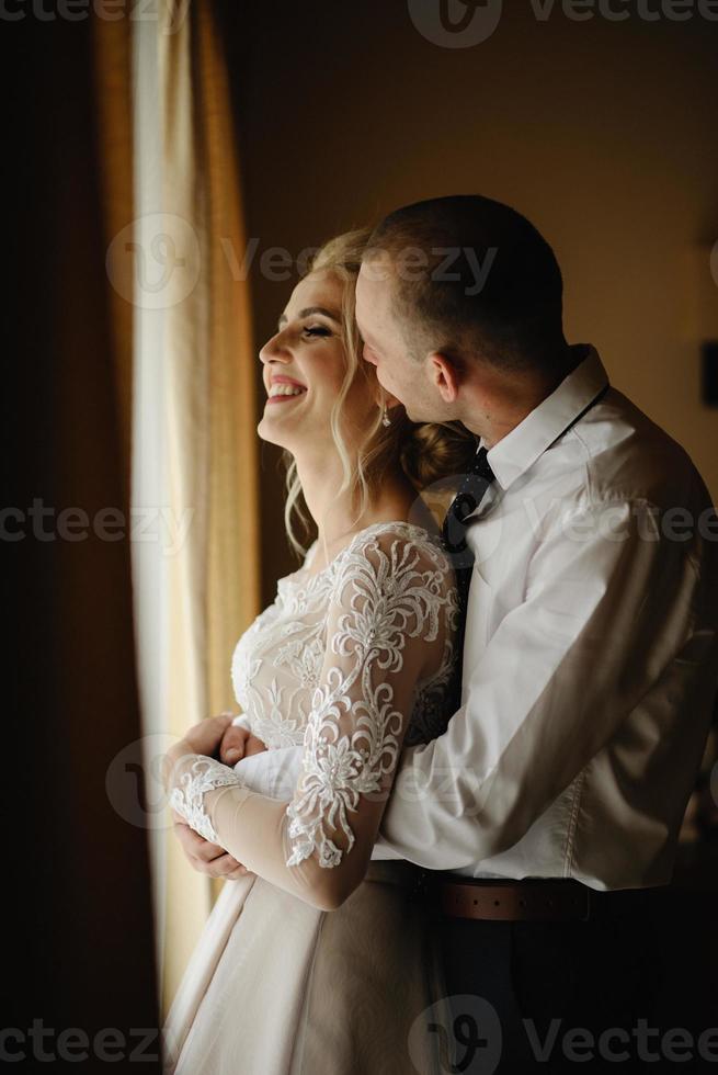magnifique mariée blonde vêtue d'une luxueuse robe blanche se prépare pour le mariage. préparatifs du matin. une femme met une robe. le marié aide la mariée. photo