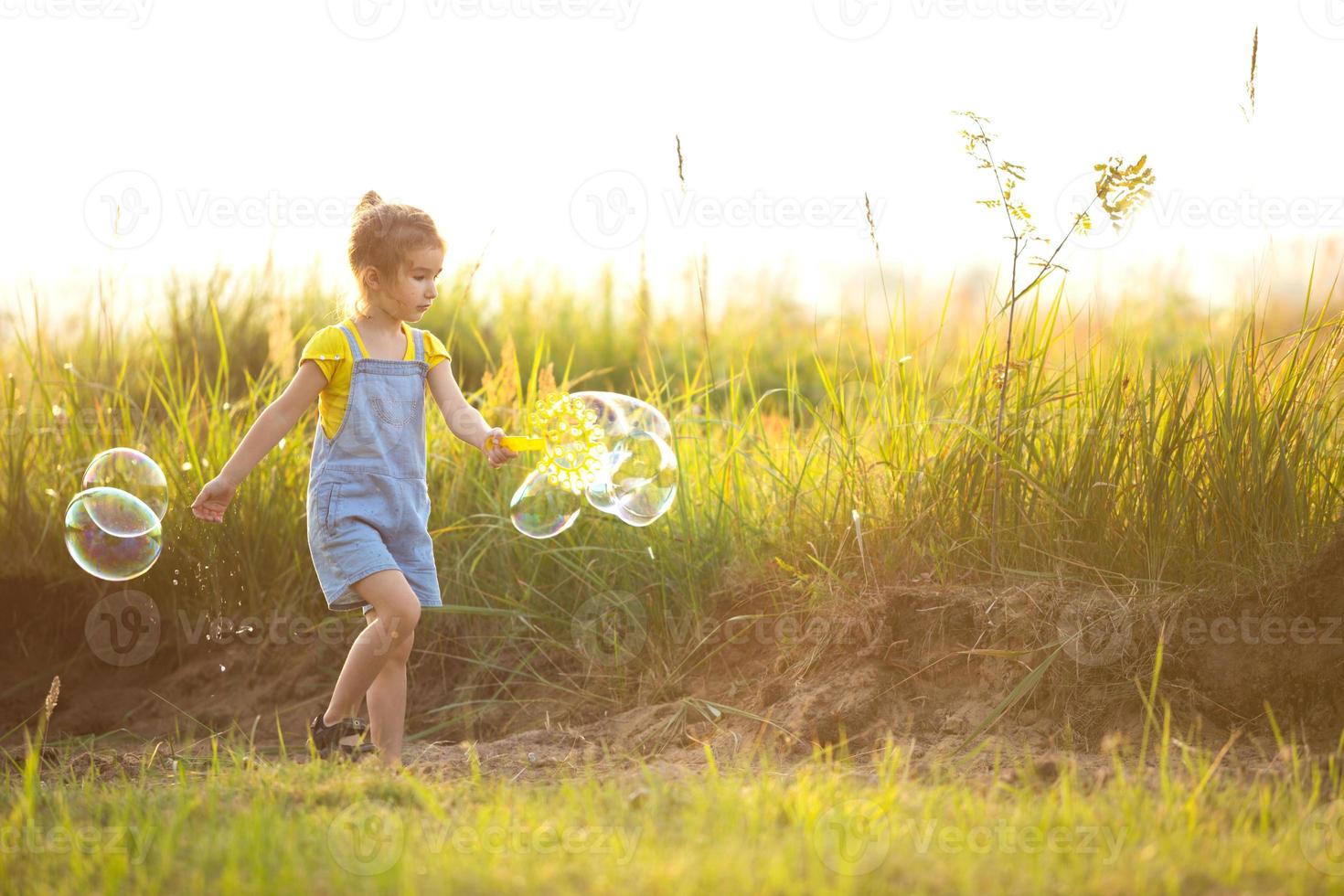 une fille en combinaison denim souffle des bulles de savon en été dans un champ au coucher du soleil. journée internationale des enfants, enfant heureux, activités de plein air. fond d'été. mode de vie sain et respectueux de l'environnement photo