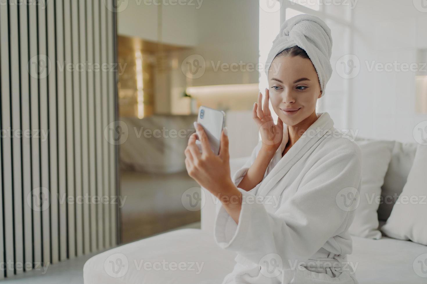 belle jeune femme appliquant une crème cosmétique tout en se relaxant sur un canapé après avoir pris une douche à la maison photo