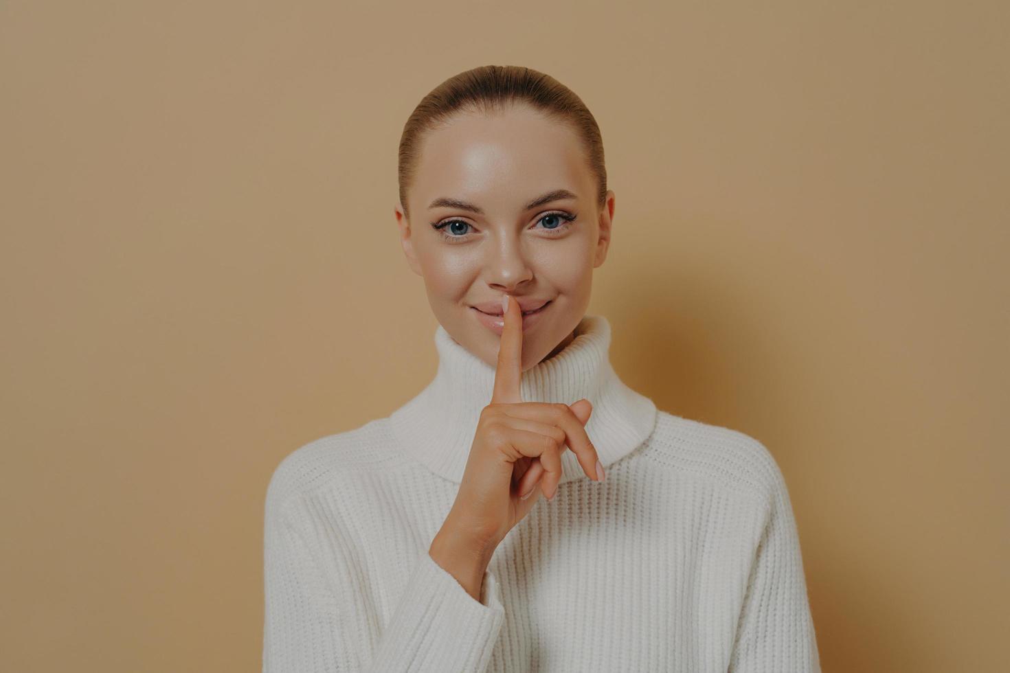 Garde ça secret. jeune femme souriante séduisante disant être tranquille avec le doigt sur les lèvres, isolée sur beige photo