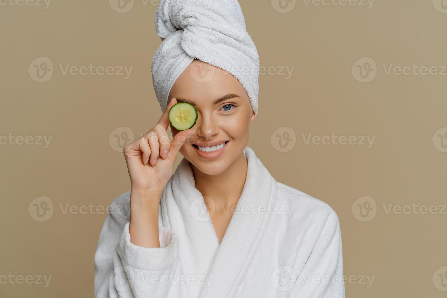 photo horizontale d'une femme ravie et rafraîchie couvre les yeux avec une tranche de concombre frais se soucie de la peau utilise des produits biologiques sourit agréablement vêtue d'une robe de bain après avoir pris une douche.