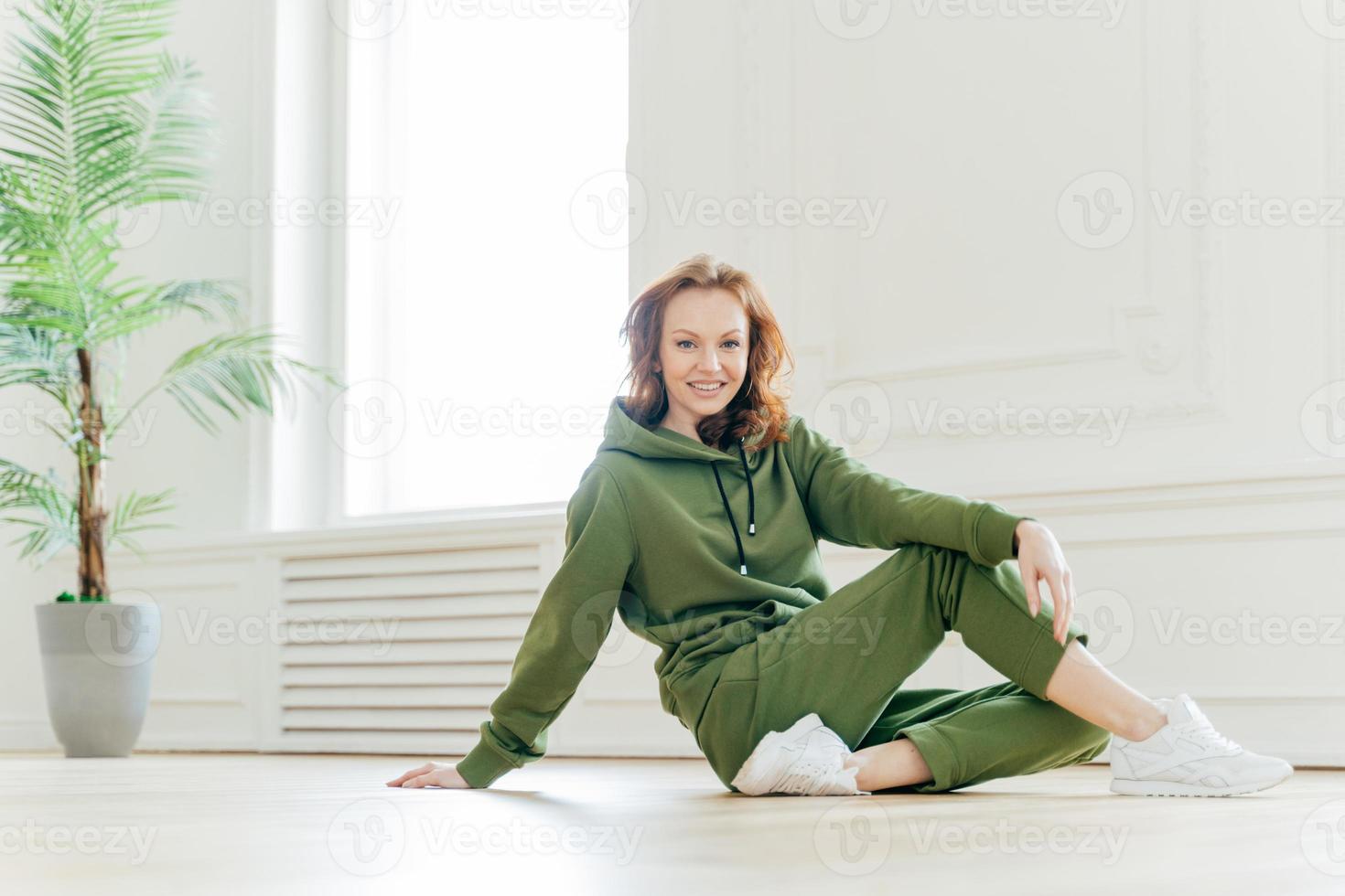 bonne forme physique femme aux cheveux rouges se penche avec la main sur le sol, a le temps de se reposer après les exercices de gymnastique, porte un survêtement vert, pose à l'intérieur, a un corps athlétique. personnes, sport, concept de style de vie photo