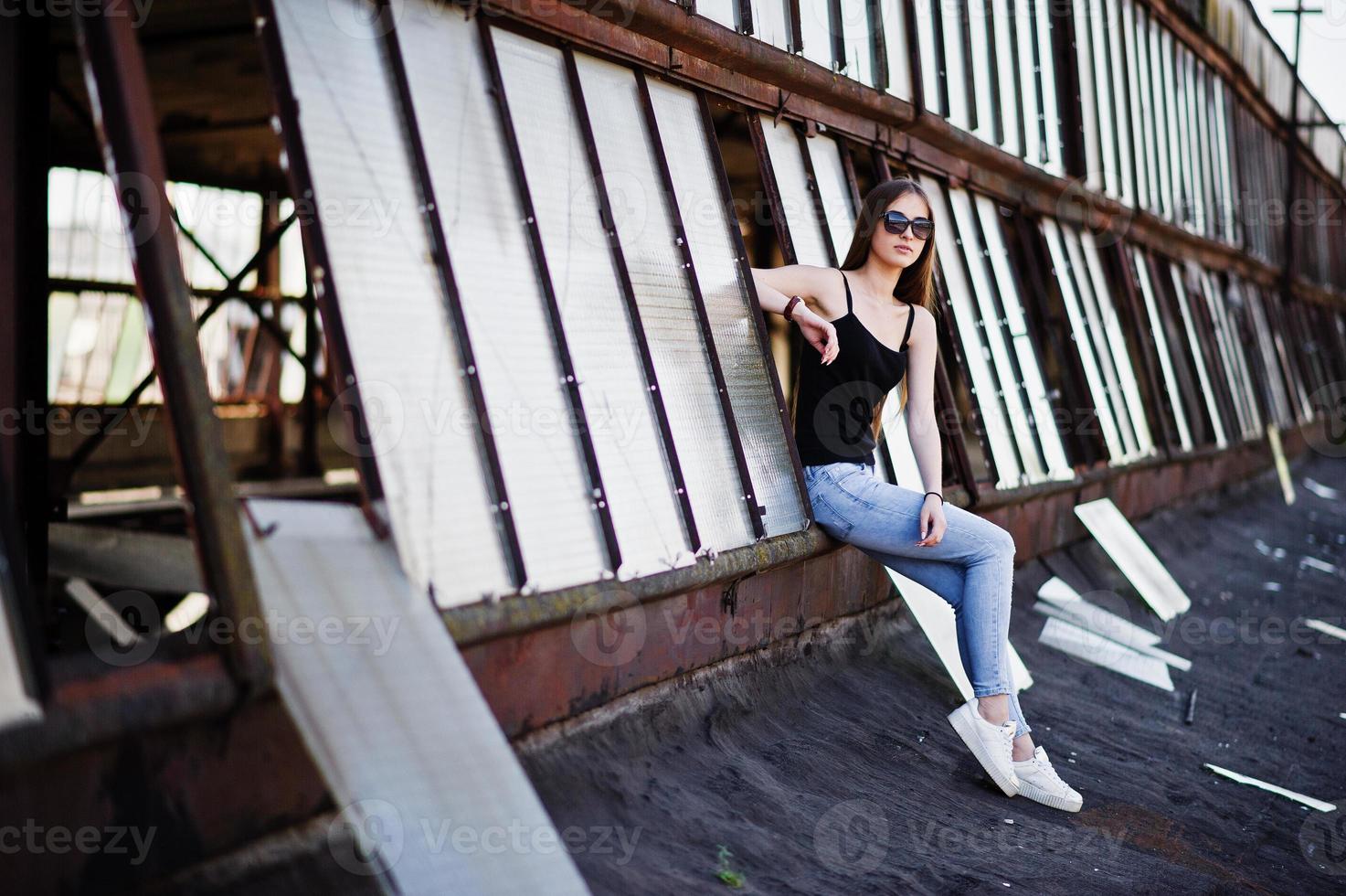 fille à lunettes de soleil et jeans posés sur le toit d'un lieu industriel abandonné avec fenêtres. photo