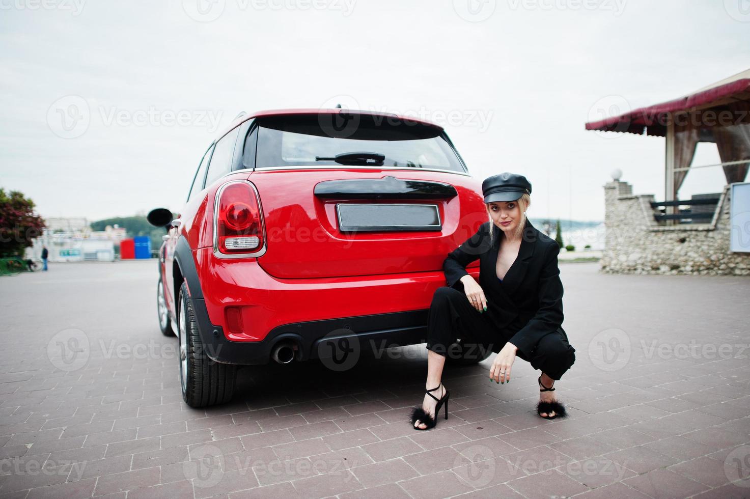 portrait d'une belle mannequin blonde sexy en casquette et en noir avec un maquillage lumineux près d'une voiture de ville rouge. photo