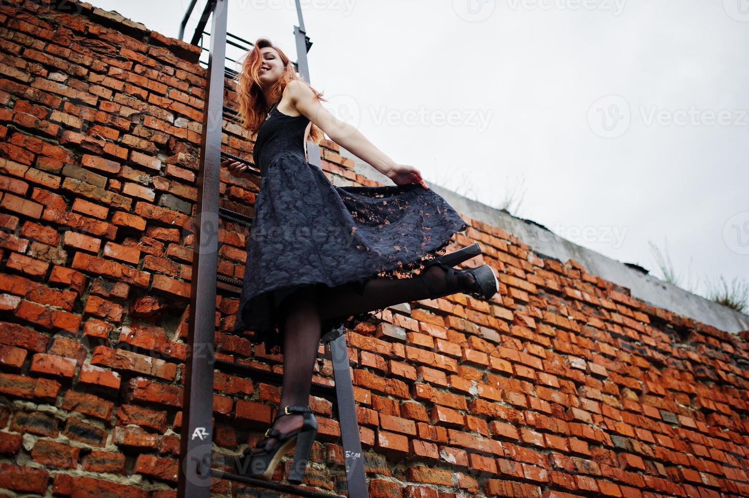 Une fille punk aux cheveux rouges porte une robe noire sur le toit contre un mur de briques avec une échelle en fer. photo