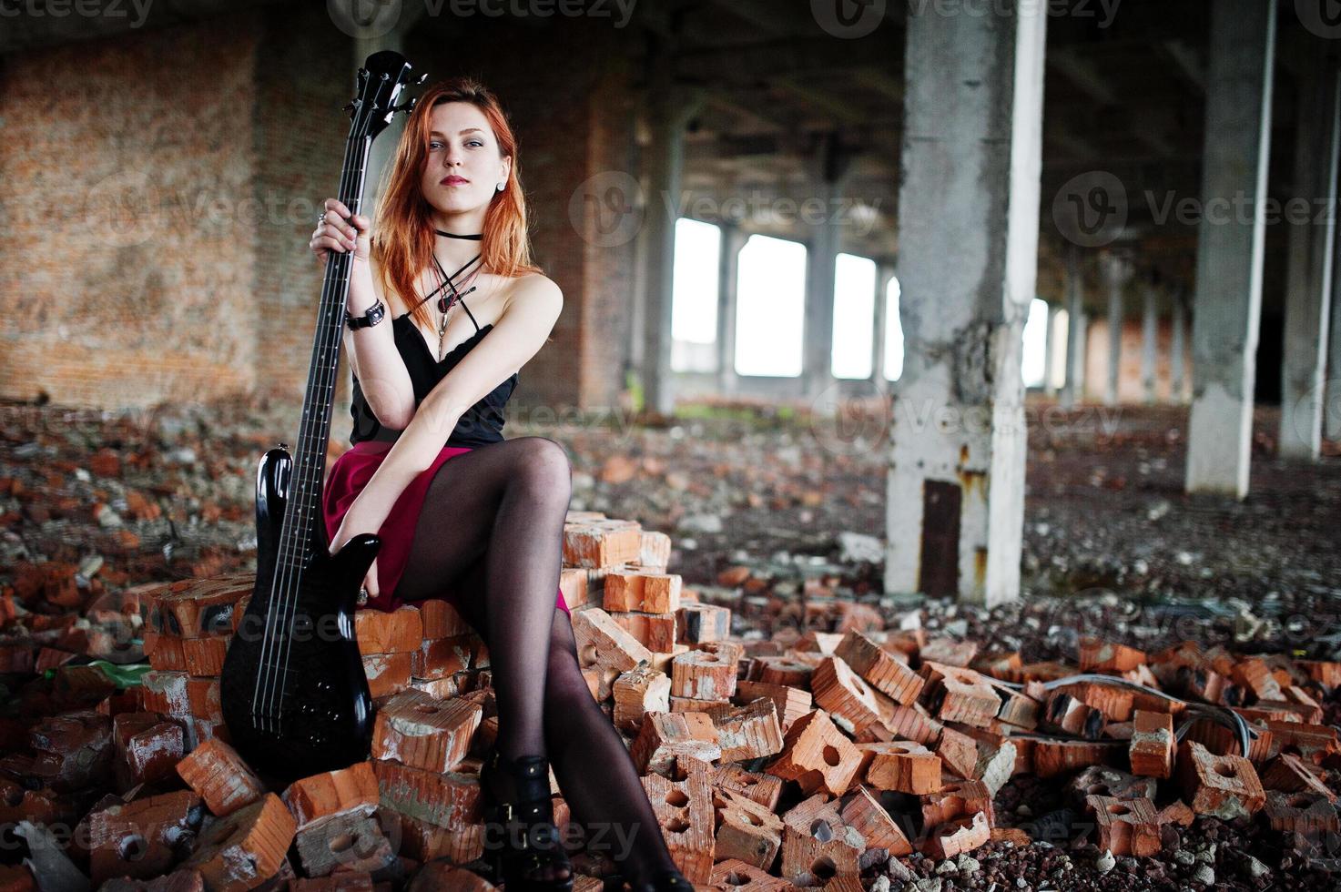 fille punk aux cheveux rouges portant une jupe noire et rouge, avec une guitare basse à un endroit abandonné. portrait de musicienne gothique. photo