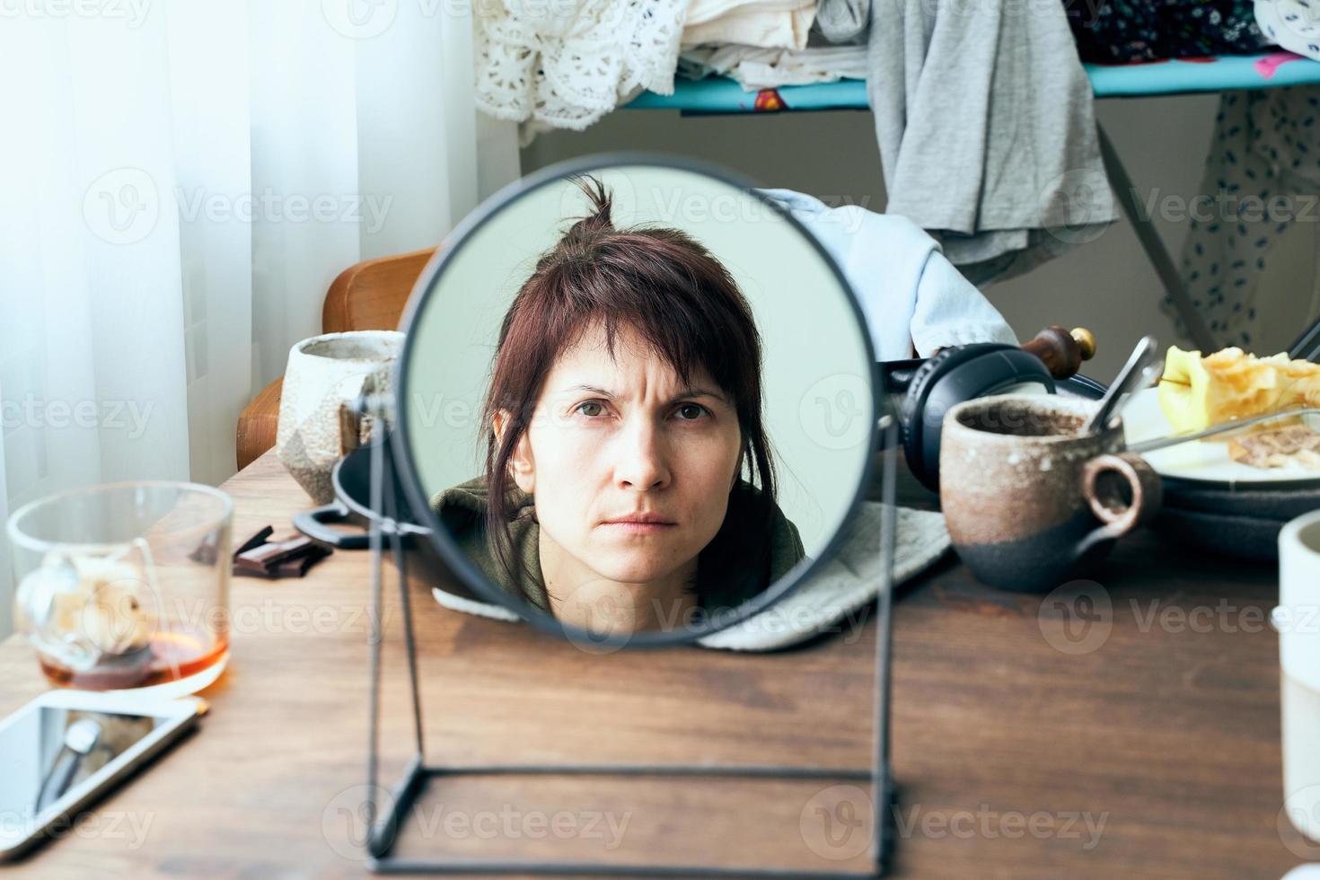 une femme se regarde dans un miroir sur fond de désordre, de vaisselle sale, de piles de vêtements photo