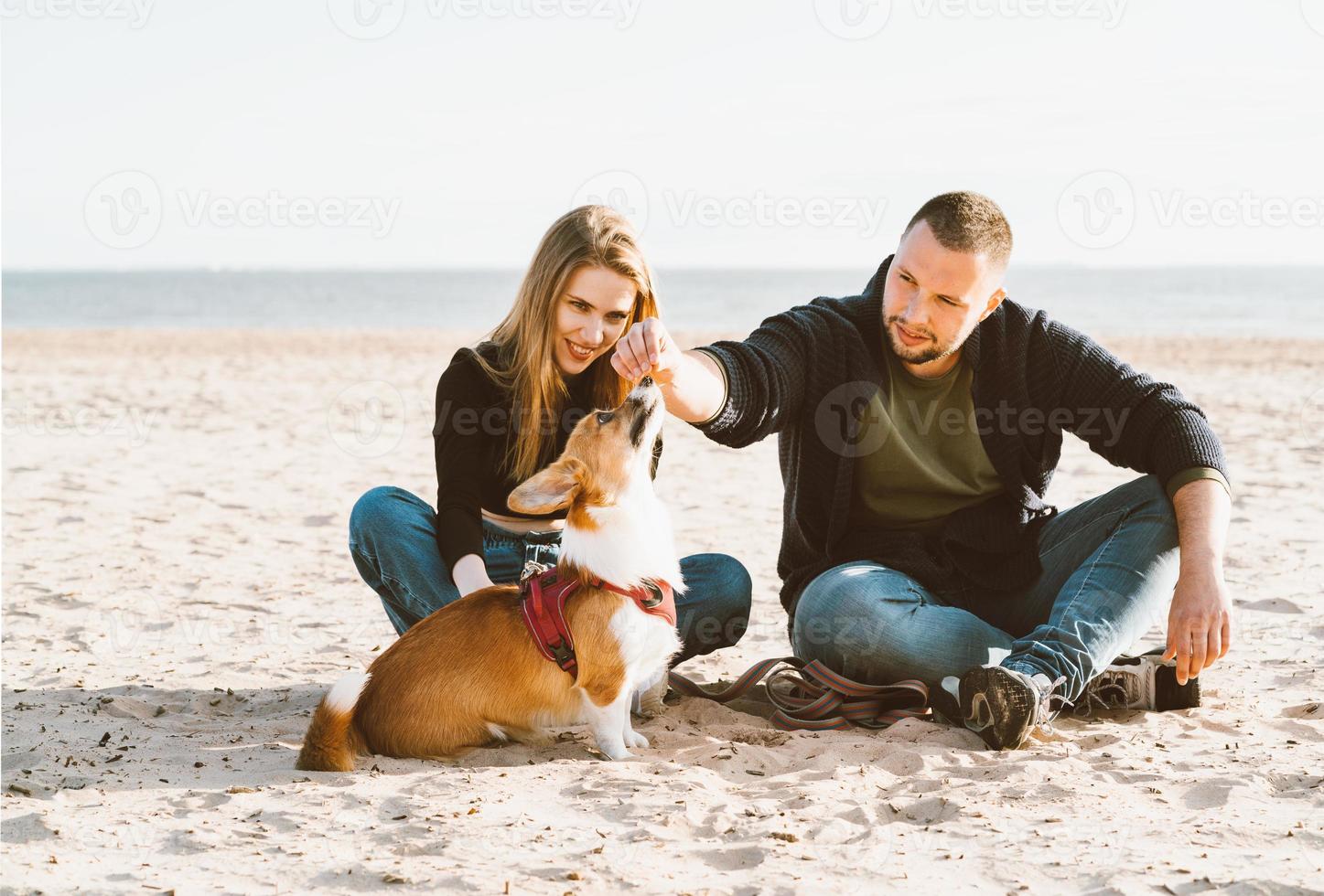 jeune couple heureux d'homme et de femme avec un chien corgi assis sur le sable. deux personnes, mâle nourrissant un animal de compagnie photo