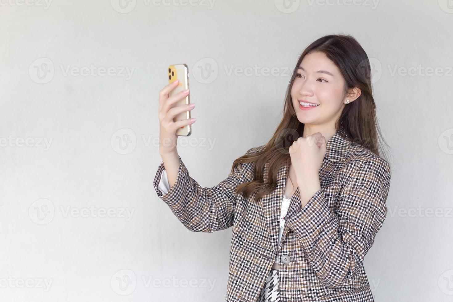 femme professionnelle asiatique aux cheveux longs noirs portant un costume à carreaux pendant qu'elle agit profiter avec succès du travail à l'aide d'un smartphone sur fond blanc. photo