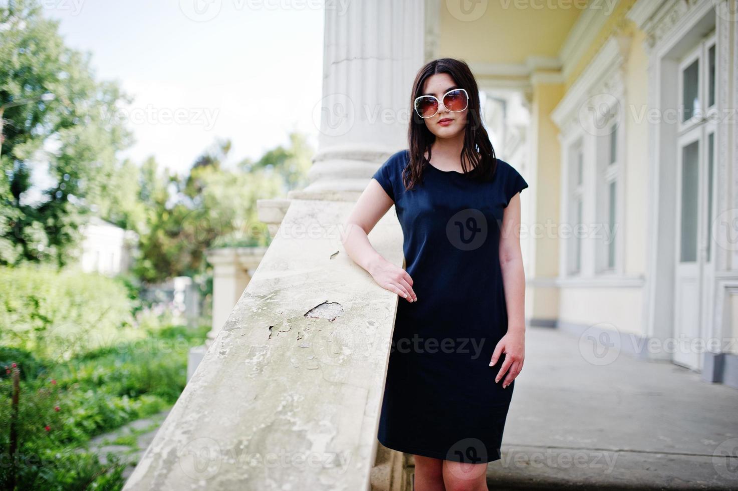 fille brune en robe noire, lunettes de soleil posées contre la vieille maison vintage, dans la rue de la ville. photo