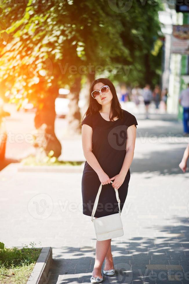 fille brune en robe noire sur lunettes de soleil avec sac à main à portée de main posant dans la rue de la ville. photo