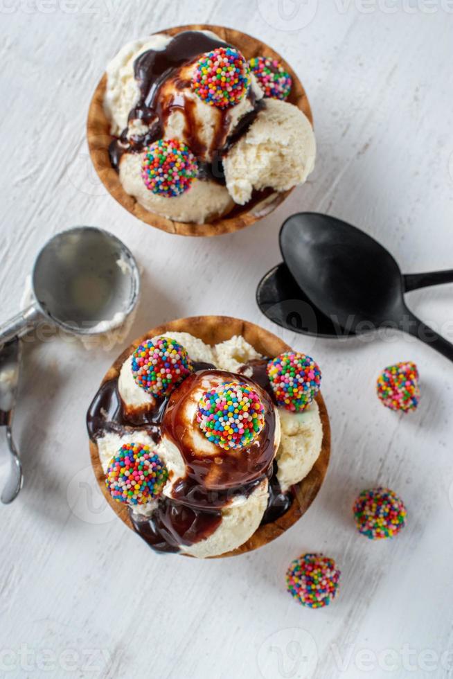 boules de crème glacée à la vanille avec des morceaux de bonbons colorés et du sirop de chocolat dans un bol à cône gaufré sur fond blanc rustique photo