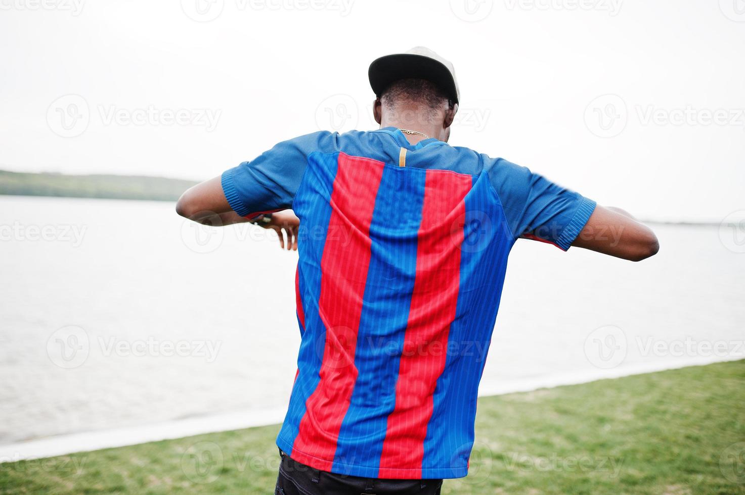 un garçon afro-américain élégant porte une casquette, un t-shirt de football montrant son dos contre le lac. portrait d'homme sportif noir. photo